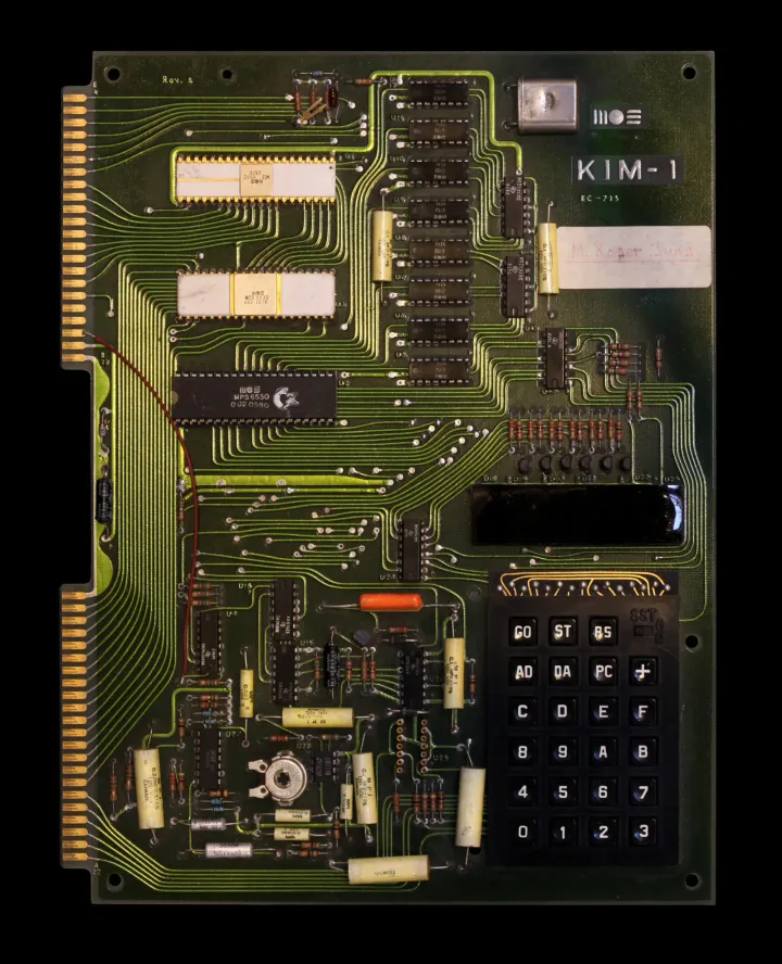 An original KIM-1 board seen from directly above on a black background: a dark green circuit board with gold edge contacts on two tabs down its left side, three 40-pin chips at the top left, a column of eight memory chips, a crystal and the KIM-1 name at the top right, the six-digit display behind a dark filter, and the black keypad of 23 keys with its SST switch at the bottom right.